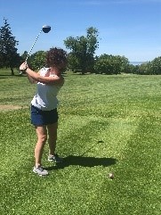 A woman with curly hair swings a golf club on a sunny day at a green golf course.