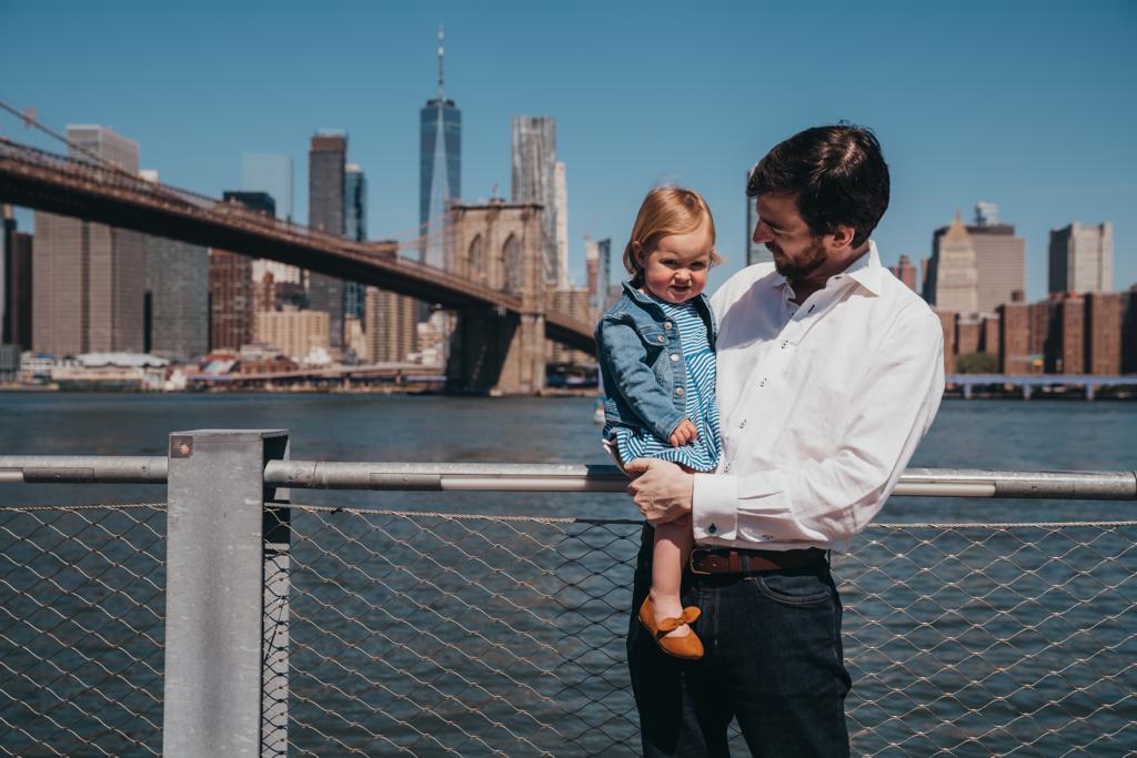 A man in a white shirt holds a child in a denim jacket by the river with the Brooklyn Bridge and city skyline in the background.