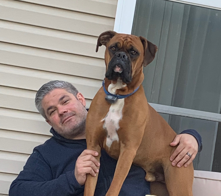 A brown Boxer dog sits on a person's lap, both are positioned near a house's wall.
