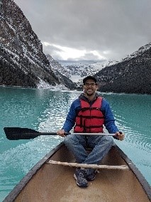A person in a blue and red life jacket sits in a canoe on serene turquoise water, surrounded by snow-capped mountains.