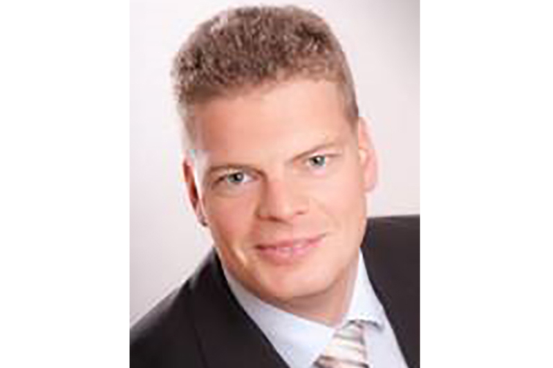 A professional man in a suit with a light blue shirt and a patterned tie, against a plain background.