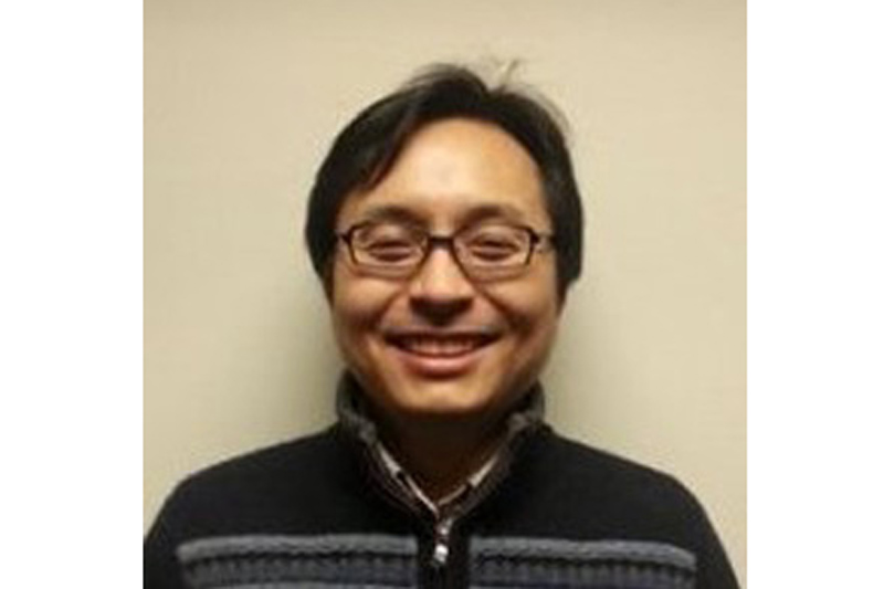 A man in a dark sweater with a patterned collar stands against a plain beige background.