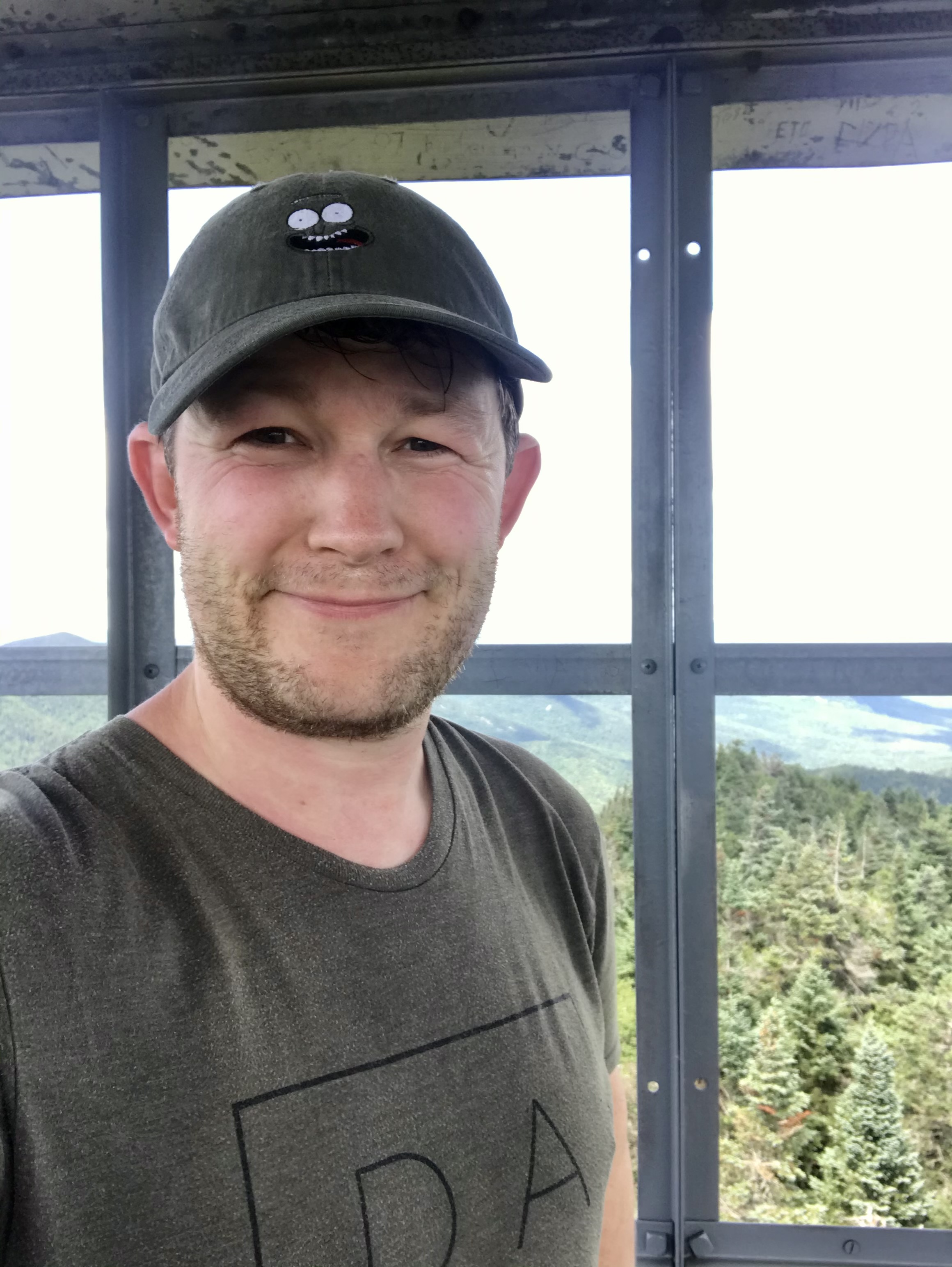 A person wearing a hat with a cartoon face stands in a lookout tower, surrounded by lush forest and mountain scenery.