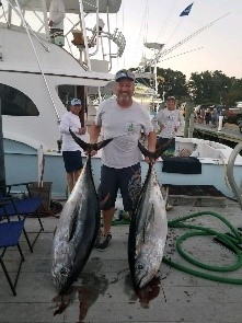 A man proudly holds two large tuna fish on a dock, with a fishing boat in the background.