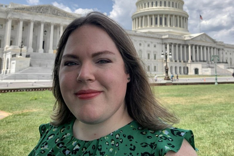 A woman in a green dress stands in front of the United States Capitol building under a blue sky.