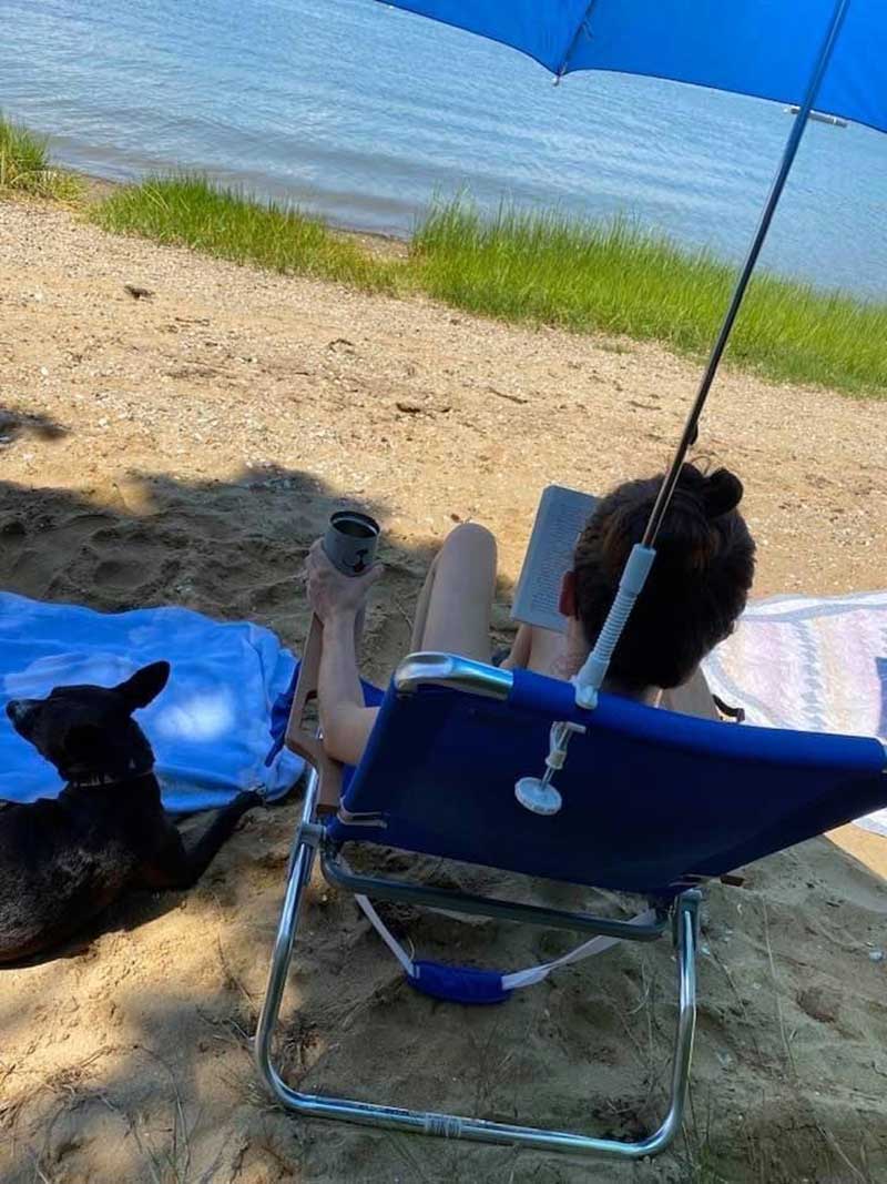 A person is relaxing on the beach under a blue umbrella, reading a book and holding a cup, with a black dog beside them.