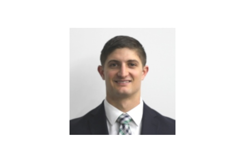 A professional man in a suit and tie stands against a plain background. He wears a patterned tie and appears confident.