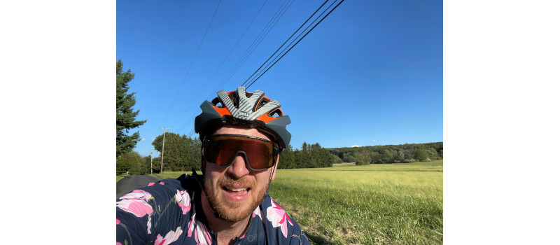 A cyclist wearing a helmet and a floral jersey poses in a sunny landscape with a clear blue sky and green fields.