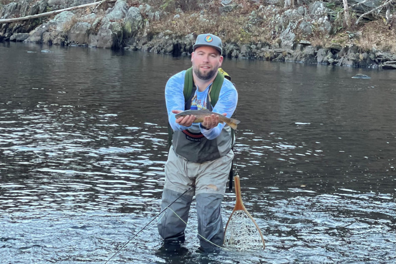 A person stands in water, holding a fish, wearing fishing gear, with a net nearby and rocky terrain in the background.