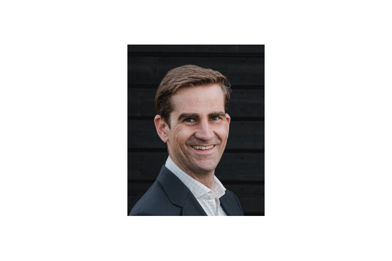 A man in a formal dark suit and white shirt is posing against a black wooden background.