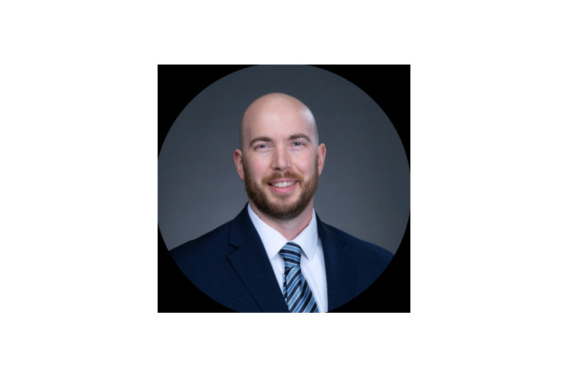 A man in a suit with a tie poses against a dark background. The image is framed in a circular crop.