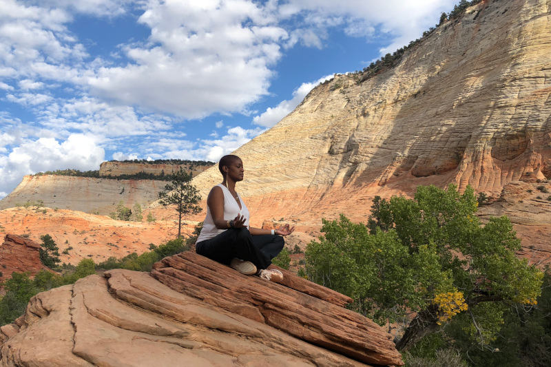 A person meditates on a rock, surrounded by vibrant red rock formations and lush greenery under a partly cloudy sky.