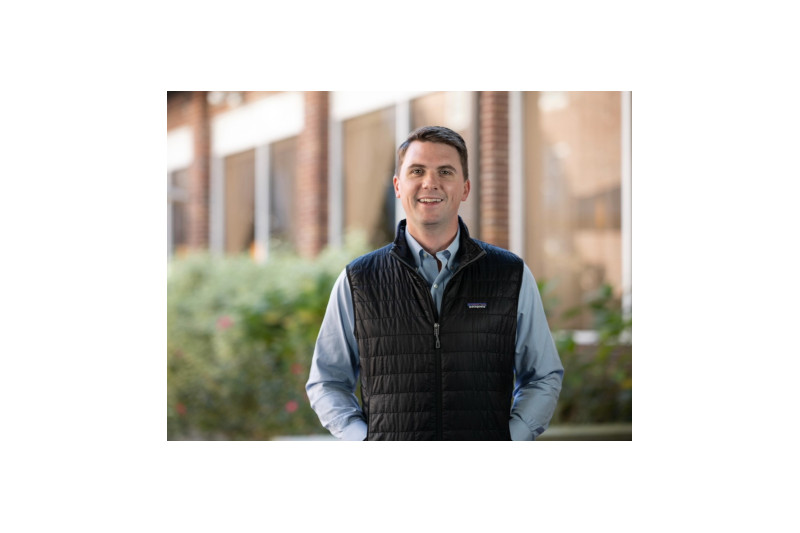 A person wearing a black vest over a blue shirt stands outdoors, surrounded by greenery and a brick building.