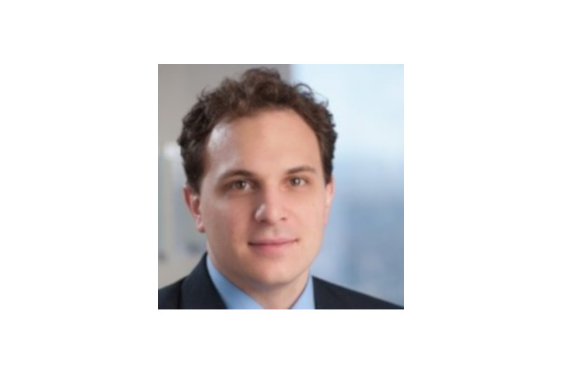 A man in a suit with a light blue shirt, posing for a professional portrait against a blurred background.