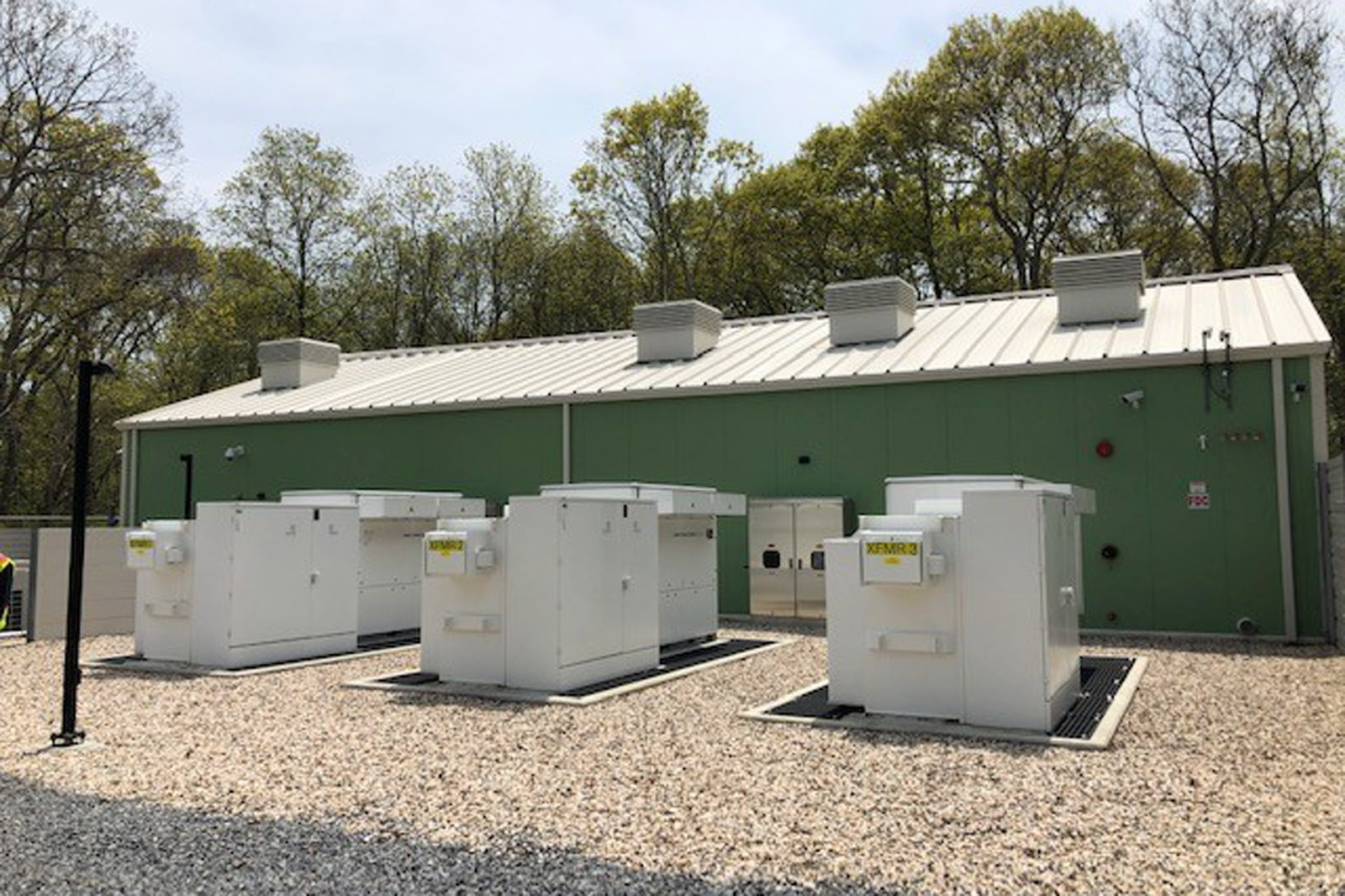 A green building with a metal roof, surrounded by white equipment containers on a gravel surface, amidst trees.