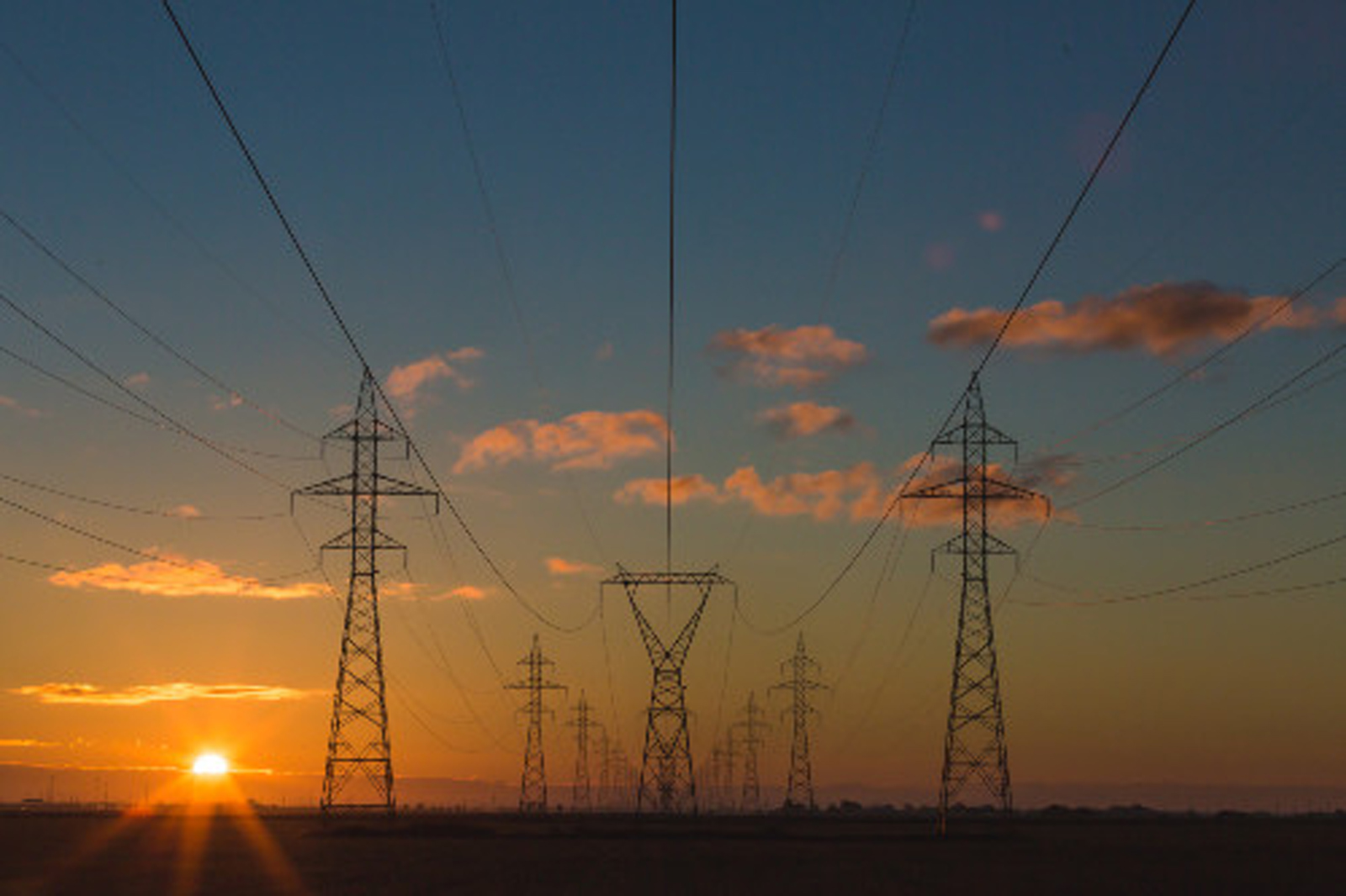 A sunset is visible behind several power lines and transmission towers silhouetted against a vibrant sky.