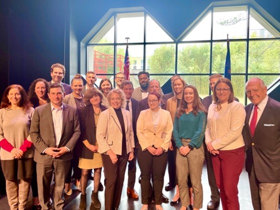 A group of people in formal attire stands together on stage, with flags and large windows in the background.