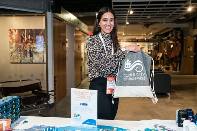 A woman presents a bag with 'COMMUNITY offshore wind' logo at an event, showcasing promotional materials on a table.