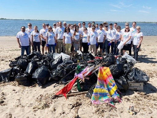 A group of volunteers gathers on the beach, posing with black garbage bags and collected debris beside a calm water body.