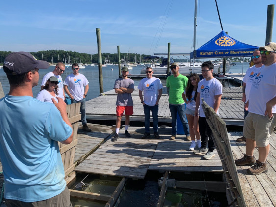 A group of people on a dock by the water, with a Rotary Club of Huntington banner and several boats in the background.
