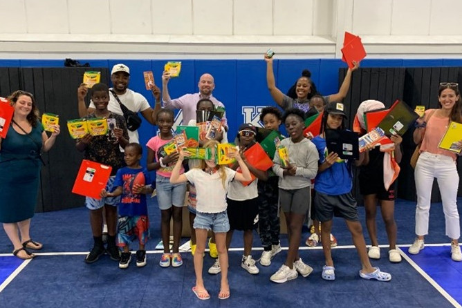 A group of children and adults holding school supplies, including crayons and notebooks, in a gymnasium setting.