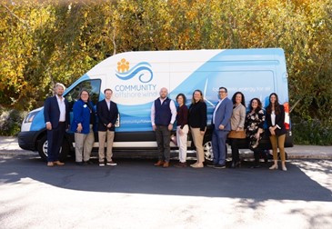 A group of diverse individuals stands in front of a blue van with 'COMMUNITY' and a wave logo, surrounded by autumn foliage.