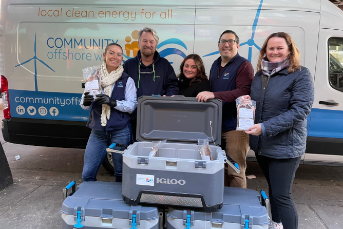 A group stands in front of a van with the logo 'COMMUNITY offshore wind', holding pamphlets and boxes of food.
