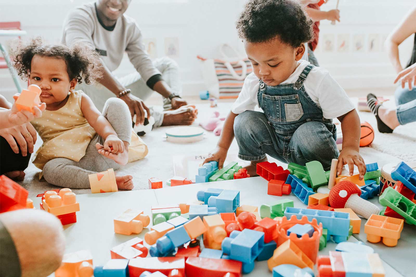 A child playing with colourful building blocks on the floor, with adults and more toys visible in a bright room.