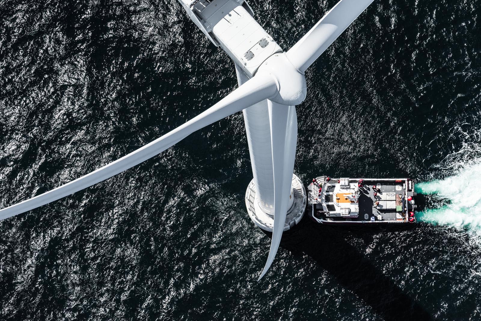 Aerial view of a large wind turbine with a boat nearby, creating ripples in the dark ocean water.