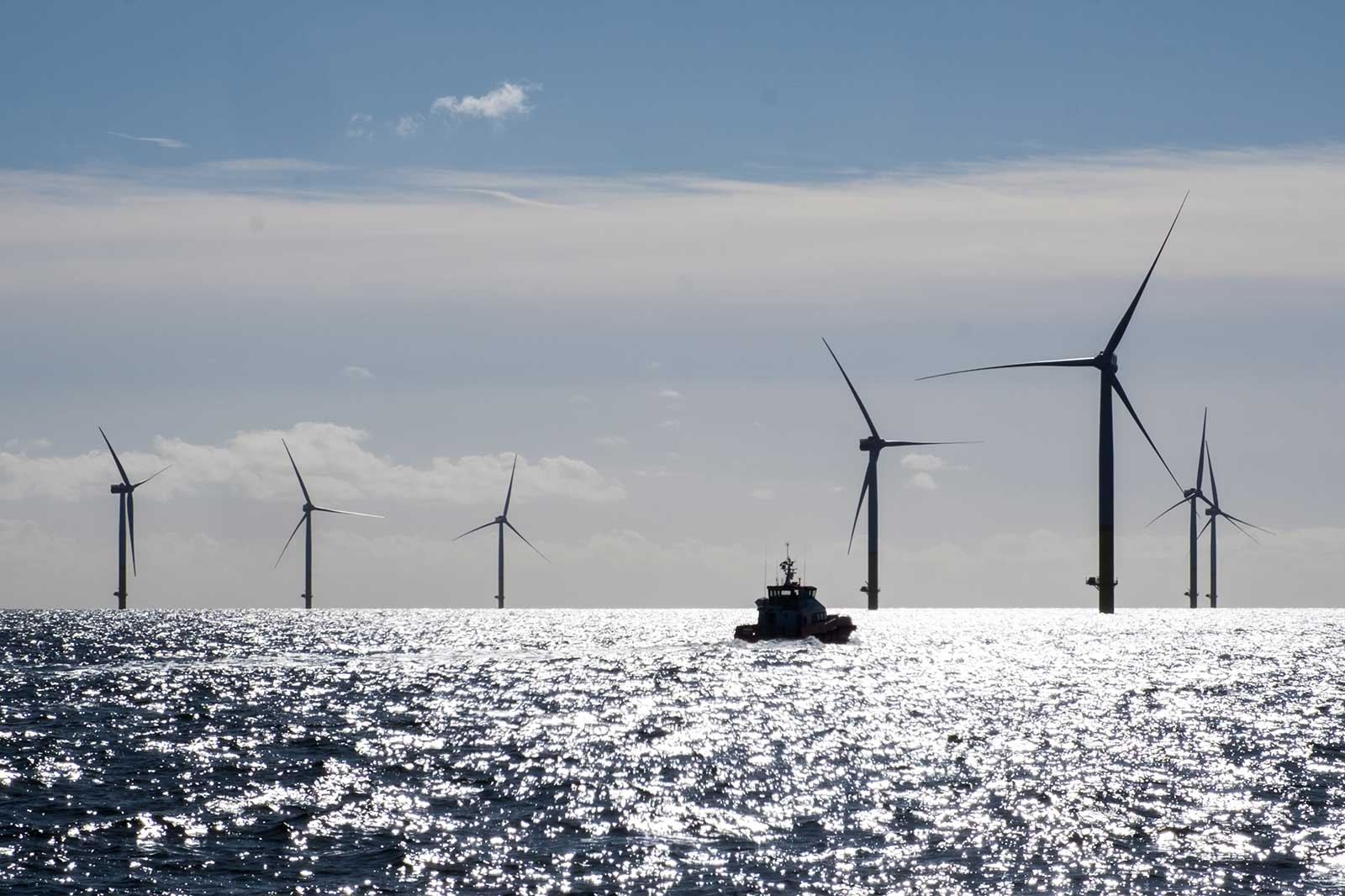 A boat sails on sunlit sea waters near a row of offshore wind turbines under a partly cloudy sky.