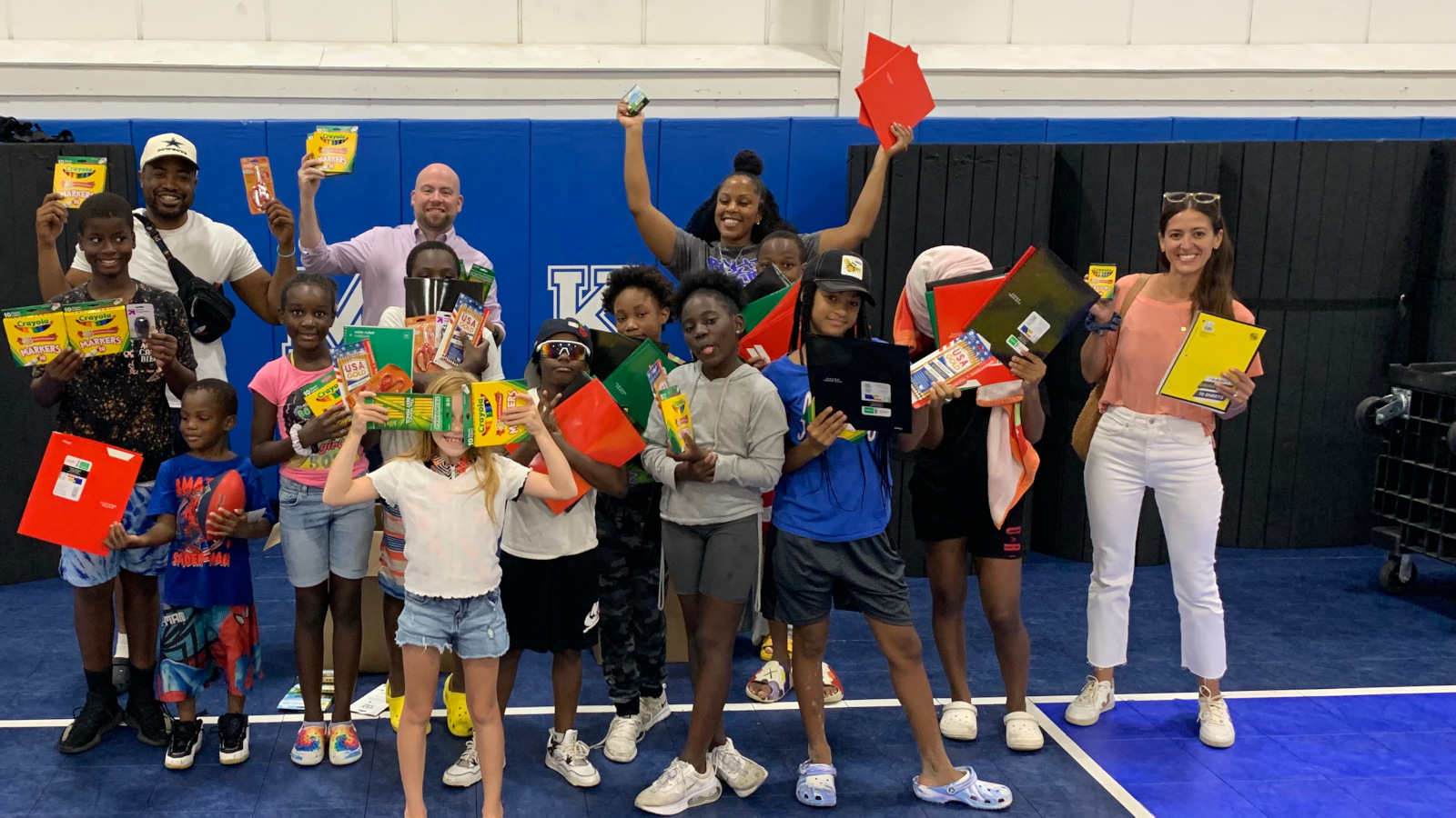 A group of children and adults holding school supplies, including notebooks and crayons, in a gym setting.