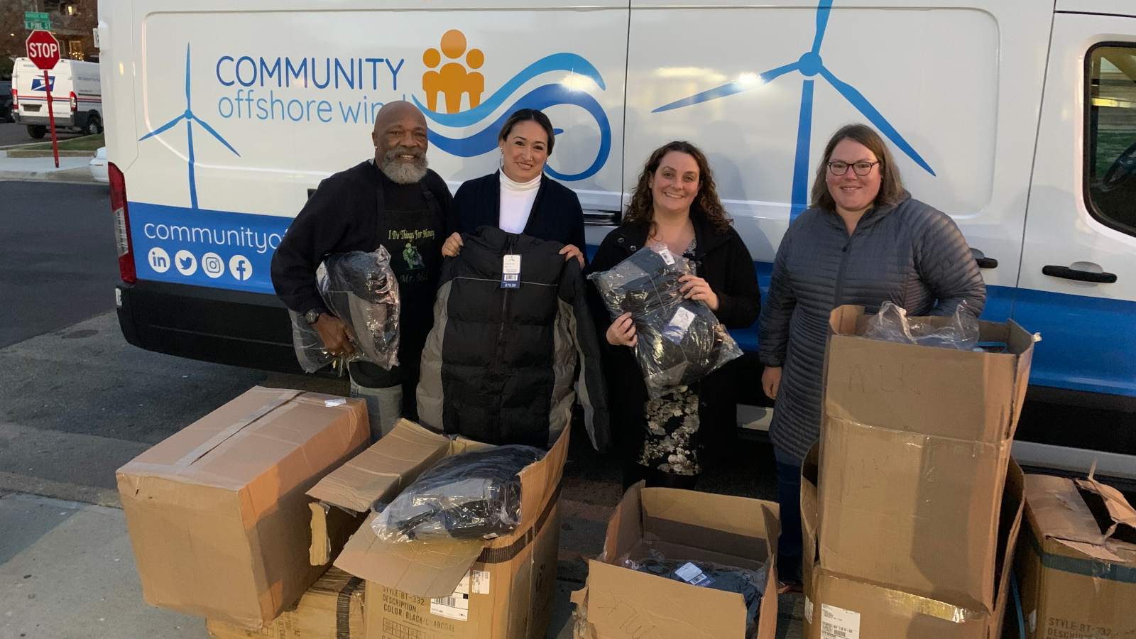 Four individuals stand in front of a van, holding jackets and surrounded by boxes of clothing donations.
