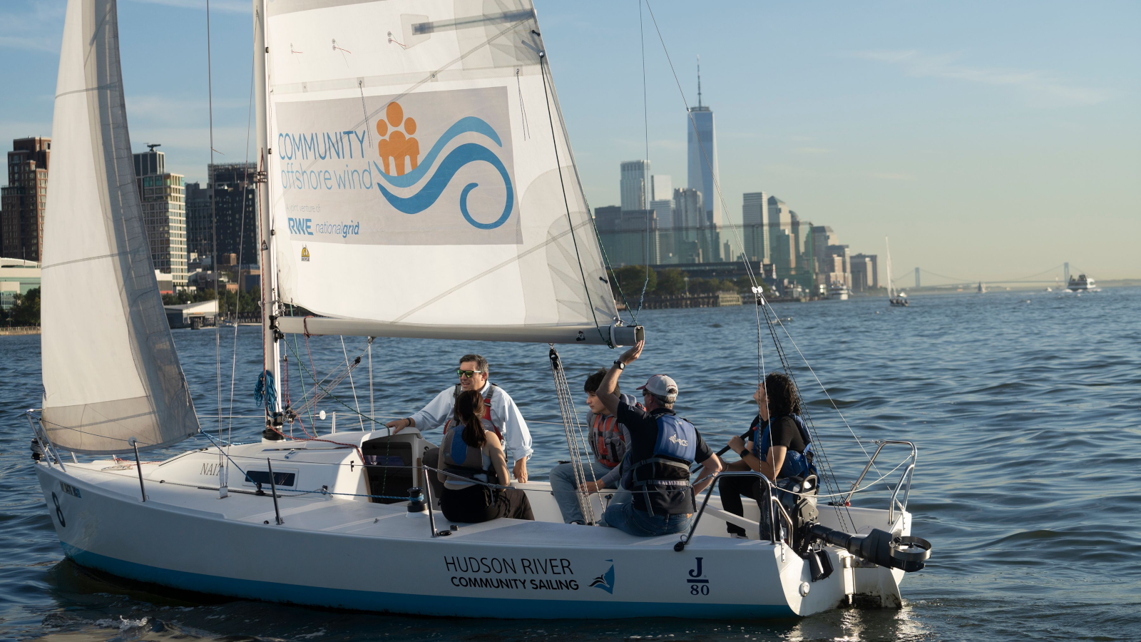 A group sailing on a small boat in the Hudson River, with the Manhattan skyline and One World Trade Center in the background.