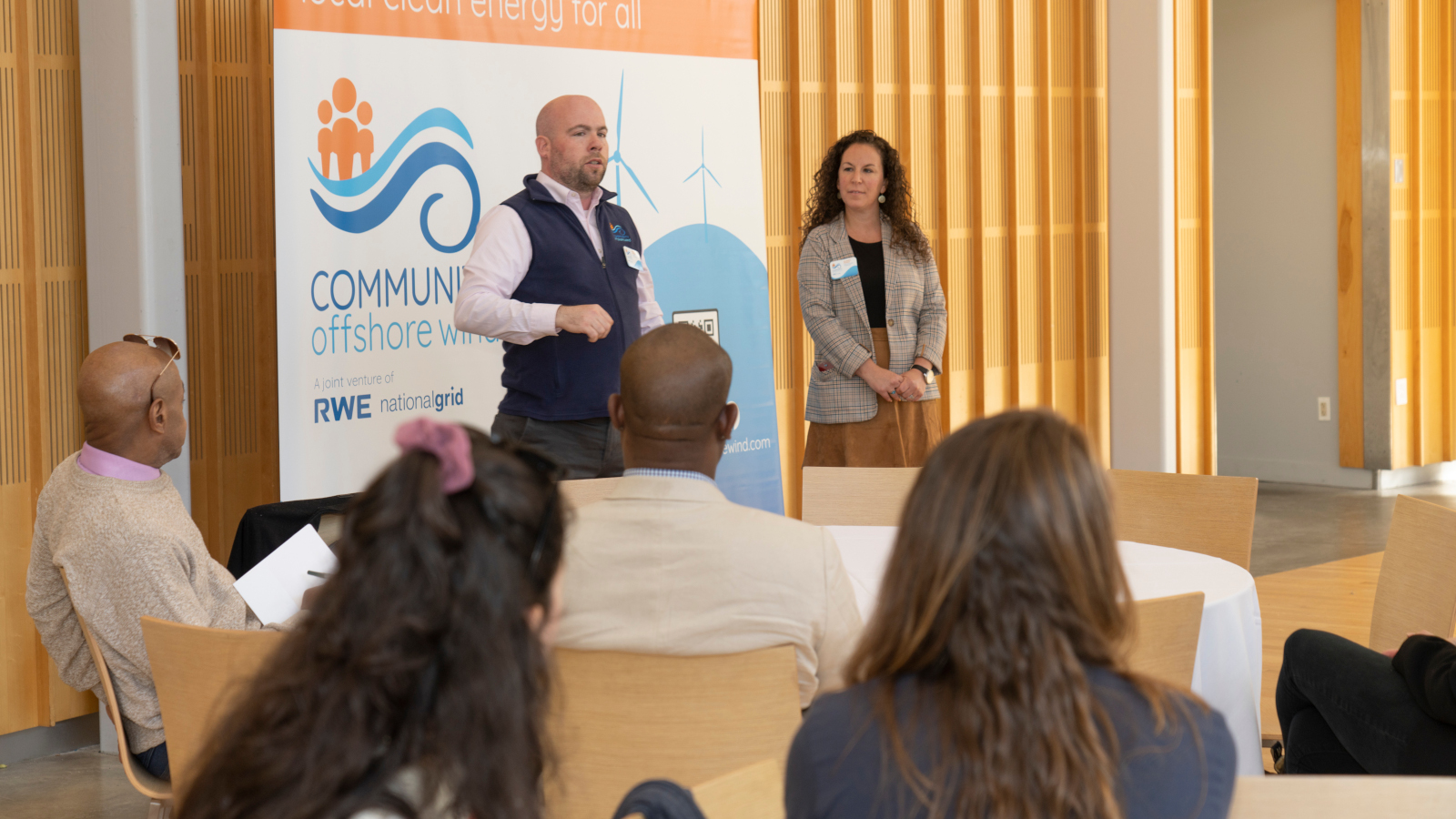 A presentation on offshore wind energy with a speaker and a woman standing beside a banner promoting community energy initiative.
