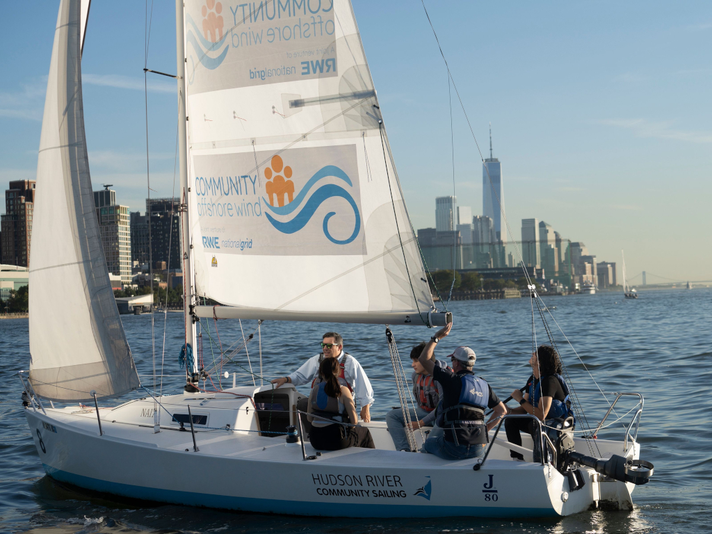 A sailboat with several people is on the water, showcasing 'HUDSON RIVER COMMUNITY SAILING' on its sail and the NYC skyline in the background.
