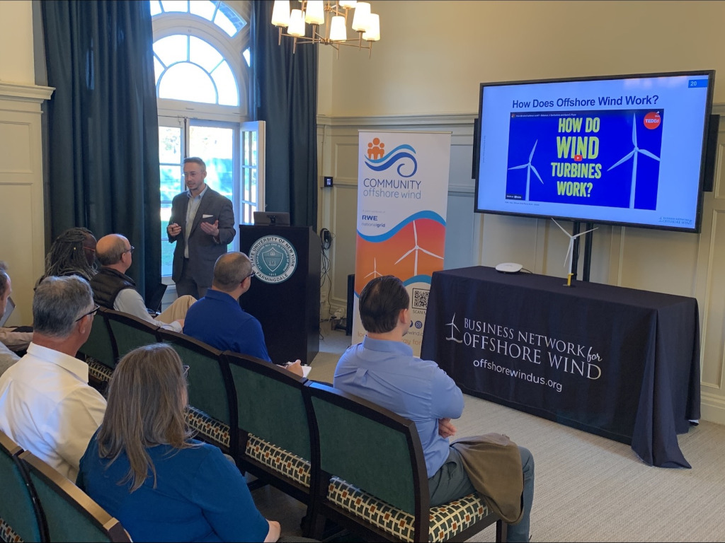 A presentation on offshore wind energy is taking place. A screen displays 'HOW DO WIND TURBINES WORK?' with an audience present.