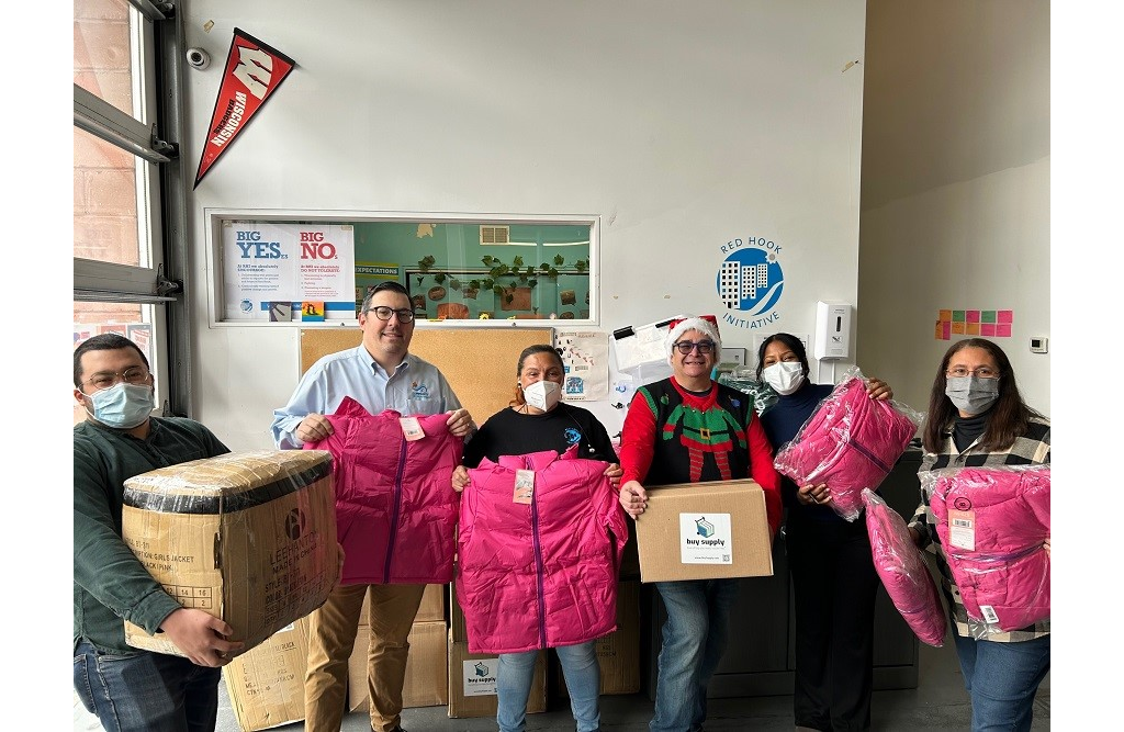 A group of six people stand in a room holding pink jackets and a donation box, smiling and wearing medical masks.