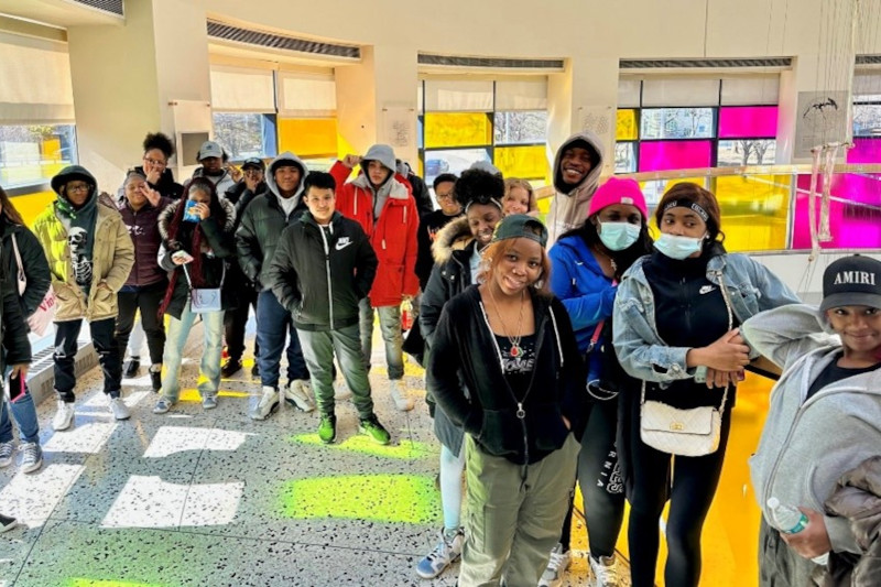 A diverse group of young people stands in a brightly lit room with colourful windows, wearing casual clothing.