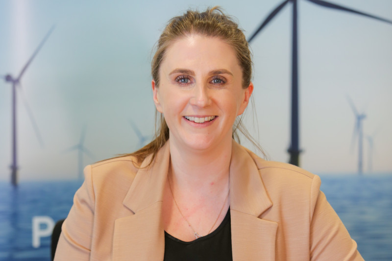 A professional setting featuring a woman wearing a beige blazer, with wind turbines visible in the background.