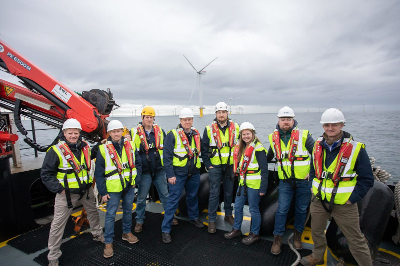 A group of ten individuals in safety gear pose on a boat with wind turbines in the background under a cloudy sky.
