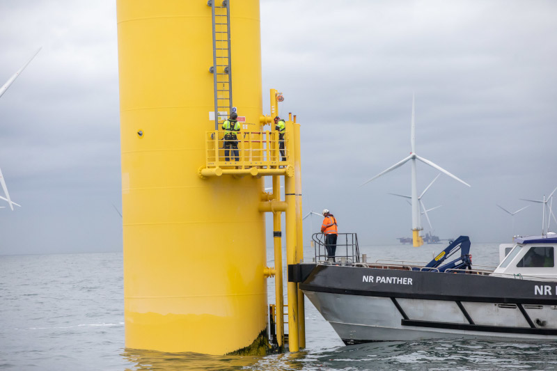 Three workers are on a yellow offshore platform, while a boat named NR PANTHER is moored nearby with wind turbines in the background.