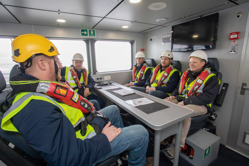 A group of six individuals in safety gear sits around a table in a modern meeting room with a TV screen.