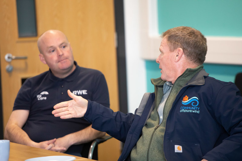 Two men are seated at a table in an office. One man gestures while wearing a Community Offshore Wind jacket.