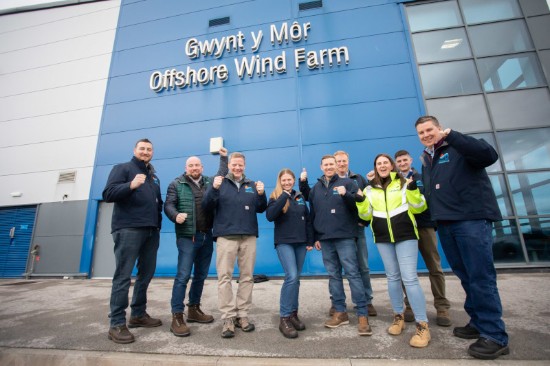 A group of nine people standing in front of the Gwynt y Môr Offshore Wind Farm building, posing happily.