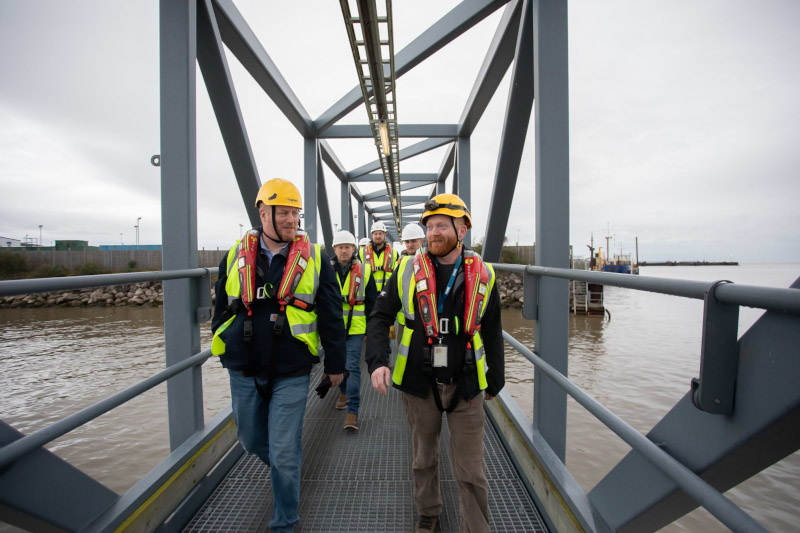 A group of workers in safety vests and helmets walks along a metal walkway over water, with boats docked in the background.