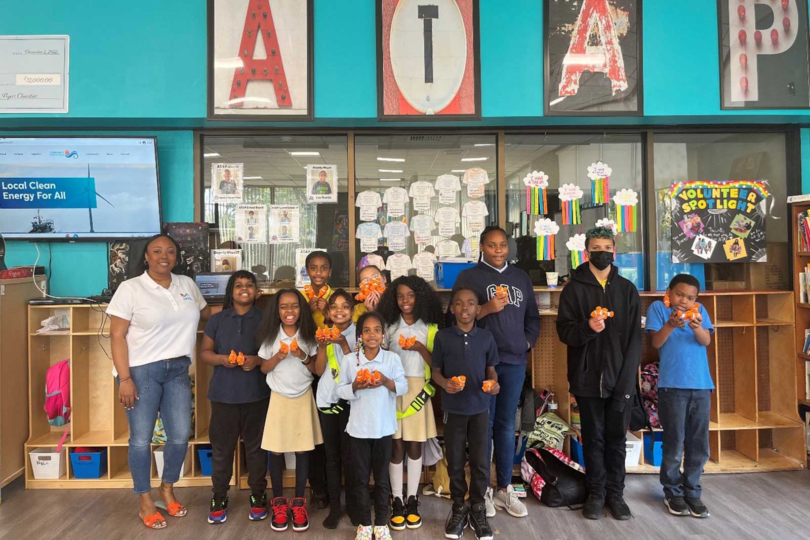 A group of children and an adult pose in a classroom, holding oranges. A sign reads 'VOLUNTEER SPOTLIGHT' on a bulletin board.