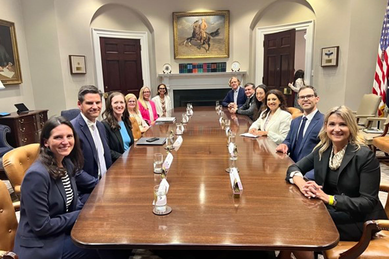 A formal meeting taking place around a long wooden table with various attendees, framed by a large horse painting.