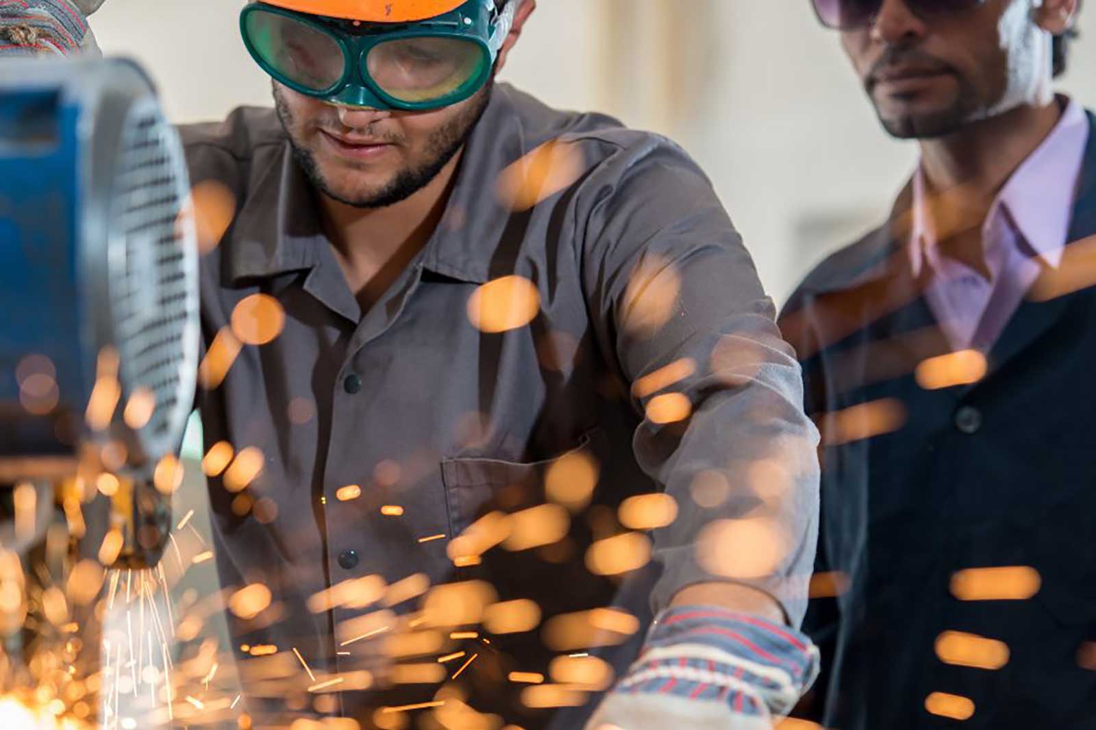 A person operates machinery that sparks, with another individual observing. Safety wear is visible, highlighting a workshop environment.