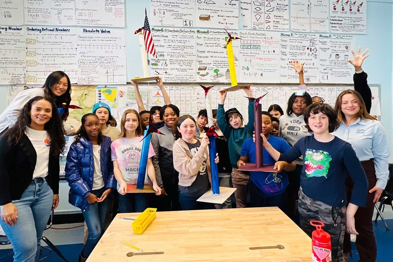 A group of children proudly display their colourful project towers in a classroom filled with educational posters.