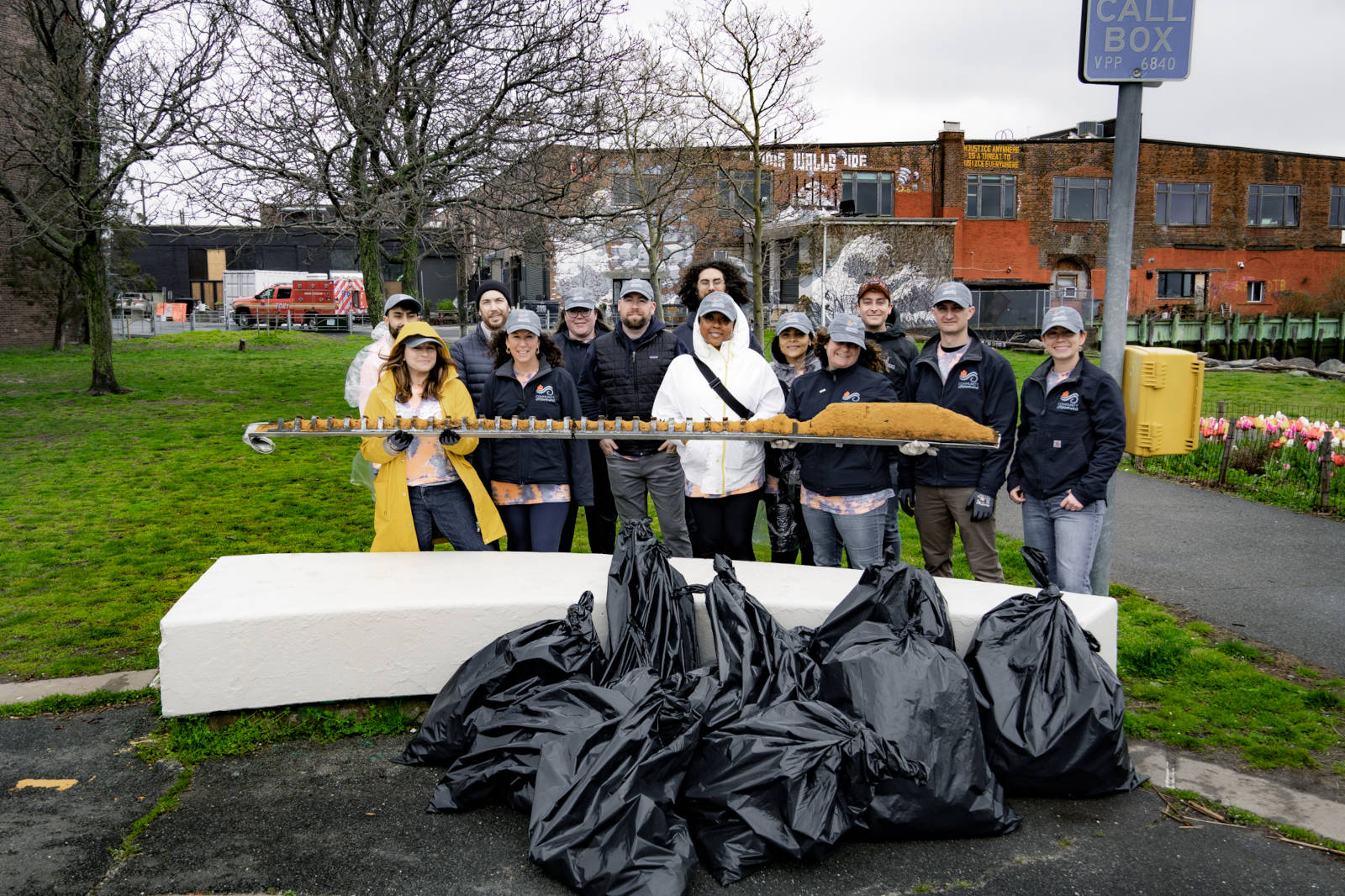 A group holds a long object while standing in front of a pile of black trash bags, with trees and a building in the background.
